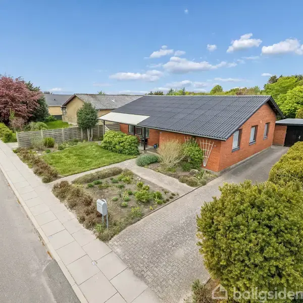 En murstensvilla med gråt tag omgivet af en velplejet have. Villaen ligger ved en rolig villavej med fortov og blomstrende træer i en forstadsmæssig bebyggelse.