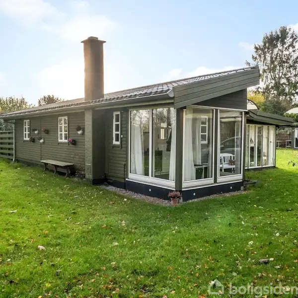 Et sommerhus står på en grøn græsplæne omgivet af træer. Huset har store vinduer og en skorsten, og det er beliggende i en landlig have.