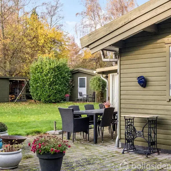 Et træhus med vinduer står ved en terrasse, hvor der er et bord og seks stole. I haven er blomsterkrukker og en lille skur omgivet af træer og buske.