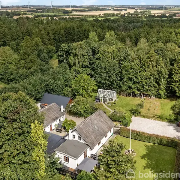 Et landhus ligger blandt frodige træer og haver. En grusvej fører til ejendommen, der har et drivhus i baggrunden. Landskabet er grønt og naturskønt.