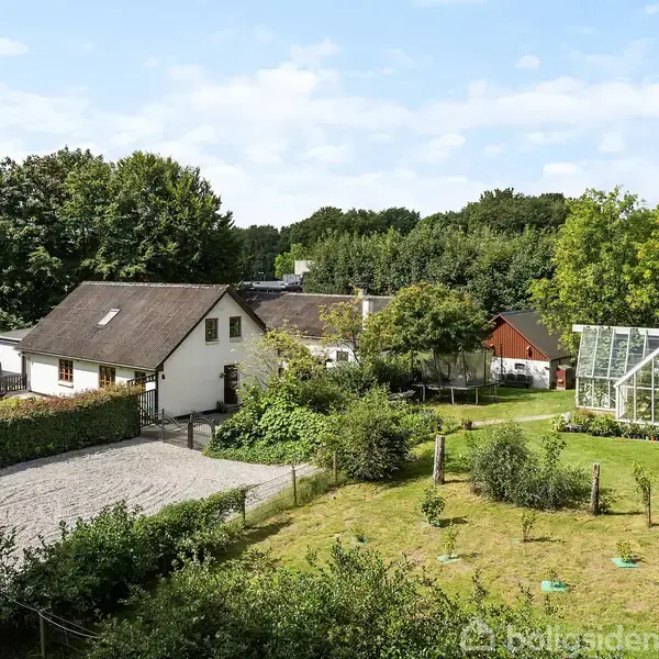 En hvid villa med skråt tag står i et grønt landskab med græsplæner og en drivhus ved siden af; omgivet af træer og buske under en blå himmel.
