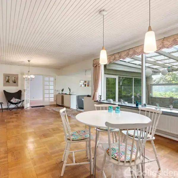Rundt spisebord med fire stole står i en lys stue. Store vinduer til venstre giver udsigt til en overdækket terrasse og have. Lysekrone hænger fra det hvide loft.