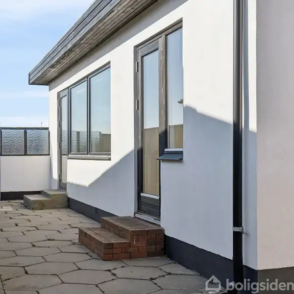 Hus med hvid facade og store vinduer, stilstand, omgivet af flisebelagt gårdhave og lavt hegn.