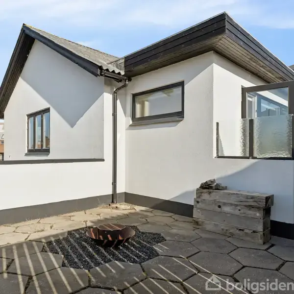 Et hvidt, moderne hus står på en flisebelagt terrasse. Vinduer og et glasparti er synlige, og der er en plantekasse nær væggen. Boliger ses i baggrunden.