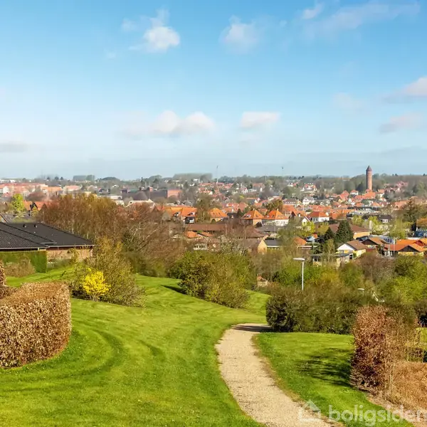 En snoet grussti fører gennem en grøn park med klippede hække. Huse og en kirketårn ses i baggrunden under en blå himmel med få skyer.