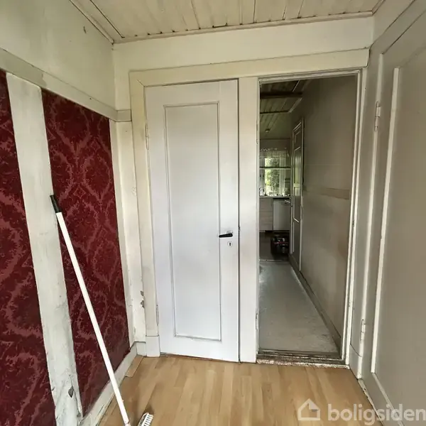 Dør står åben mod en smal gang med udsigt til et andet rum i baggrunden. Væggene er rødt tapet, og gulvet er i lyst træ.