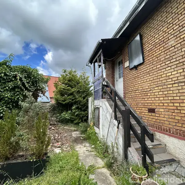 Et ældre murstenshus med en udvendig trappe fører op til en veranda. Omgivet af en have med buske og træer, under en overskyet himmel.
