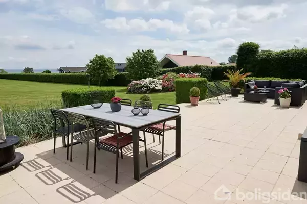 Havebord med stole står på en flisebelagt terrasse. Haven er omgivet af grønne buske, blomster og græsplæner med udsigt til et hus og himlen i baggrunden.