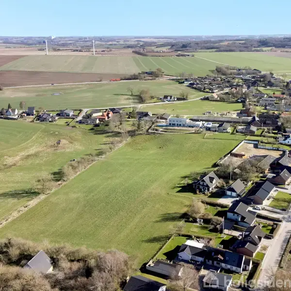 Landsby vises med række af huse langs veje, omgivet af åbne marker og landbrugsjord. Distant vindmøller ses i baggrunden på det åbne, landlige landskab.