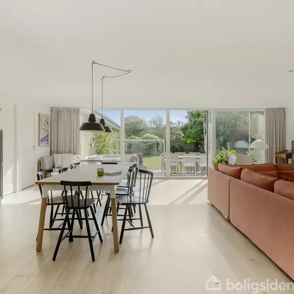 Stue med lyserød sofa og spisebord med sortmalede stole. Sollys strømmer ind gennem store vinduer, der åbner ud til en terrasse med udsigt til en grøn have.