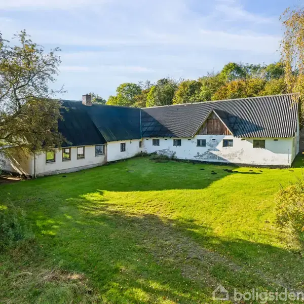 Et hvidt hus med skråtag står stille omkranset af en stor, grøn græsplæne og træer. Himlen er klar blå i baggrunden, hvilket indikerer solrigt vejr.
