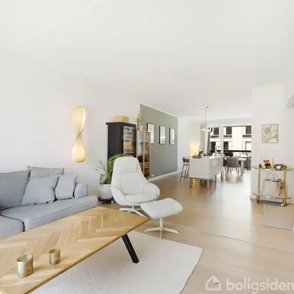 En lys stue med en grå sofa, et sofabord og en hvid stol. Der er en spiseplads i baggrunden og moderne indretning med vægkunst og belysning.