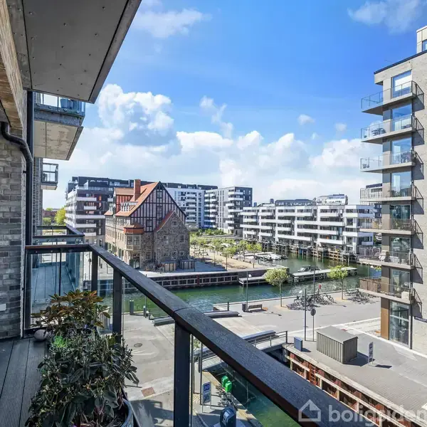 En altan med glasrækværk fyldt med planter. Den har udsigt til moderne lejlighedsbygninger og en kanal på en solrig dag. Urban miljø med klar blå himmel i baggrunden.