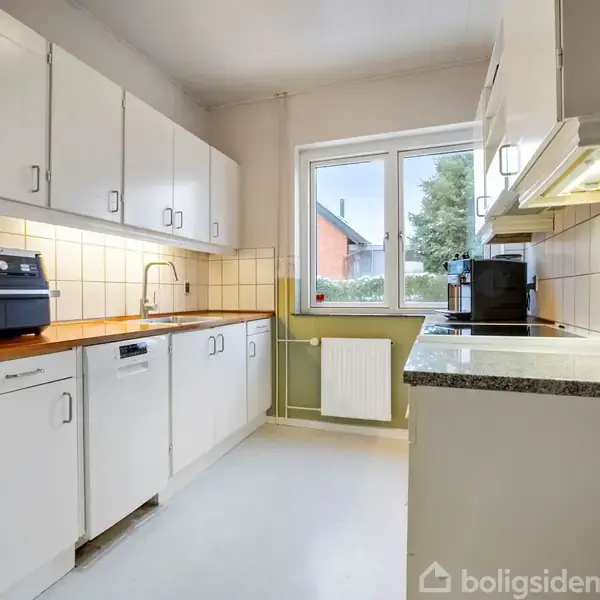 Et køkken med hvide skabe, en brødrister på bordpladen og en stor vindue i baggrunden, der viser en træfyldt udendørsscene. Radiator under vinduet.