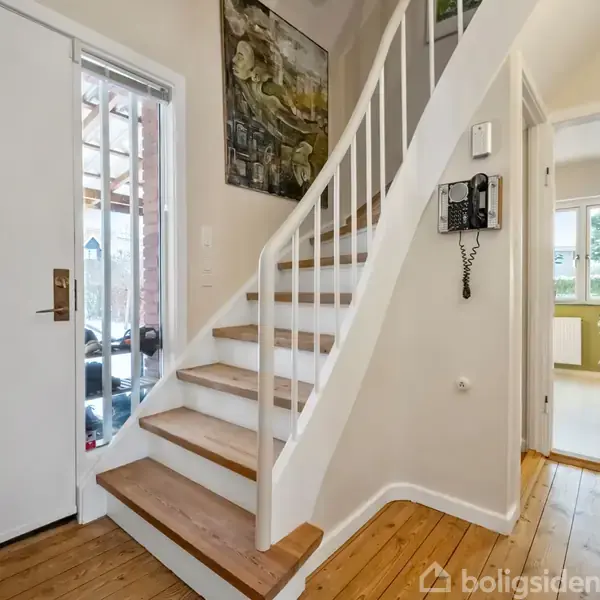 Trappe fører opad i en lys entre med træplanker på gulvet, indrammet af en åben dør til venstre. Et maleri hænger på væggen, og der ses en telefon.