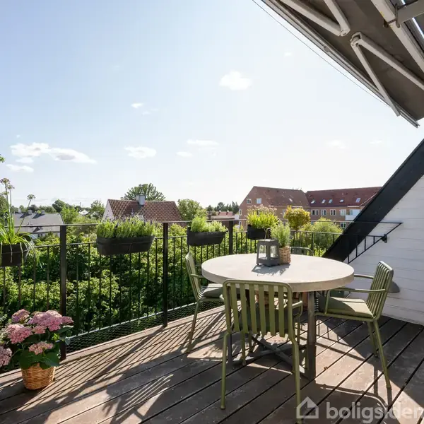En rund havebord med stole står på en træterrasse. En lanterne står på bordet, omgivet af planter i krukker. Terrasse har udsigt over grønne haver og hustage under en blå himmel.
