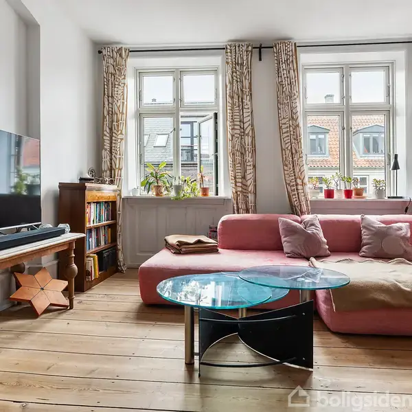 En lyserød sofa står foran store vinduer med gardiner. Rundt glasbord er placeret foran sofaen. Stuen har en bogreol og et TV på et træbord. Planter i vindueskarmen.
