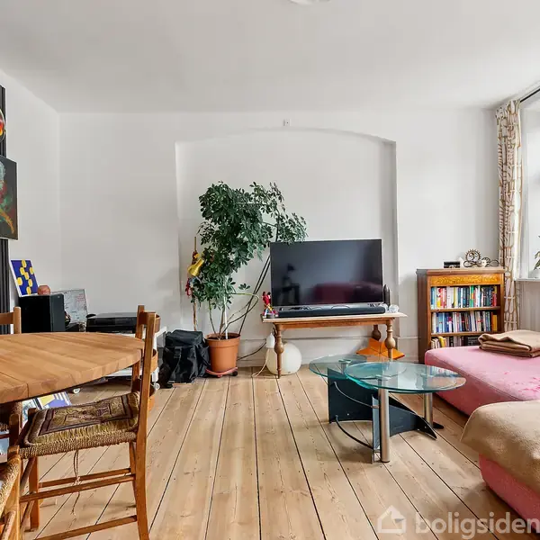 Stue med lyserød sofa ved vinduet, tv på væggen med planter ved siden af, spisebord med stole, kunst på væggen og en bogreol til højre.