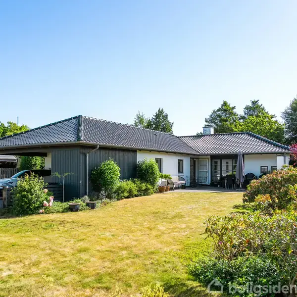 En etplans villa med sort tegltag og hvide vægge står i en velplejet have. En carport er til venstre, omgivet af buske og træer under en klar himmel.