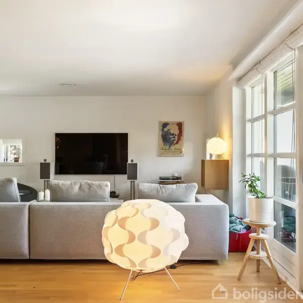 En moderne stue med en stor grå sofa vendt mod et vægmonteret TV. En unik lampe står på gulvet, og en skydedør fører ud til en terrasse. En lille plante står på en taburet.