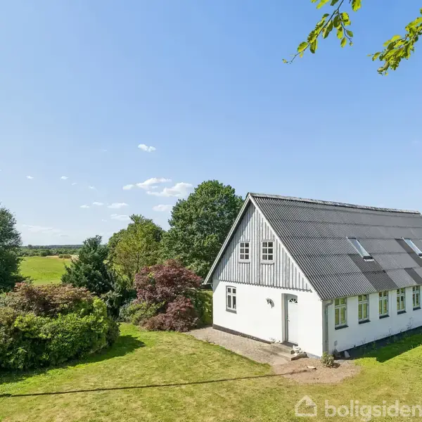 Et hvidt hus med skråt tag står i en grøn have. Træer og buske omgiver huset, og en høj flagstang er placeret til venstre.
