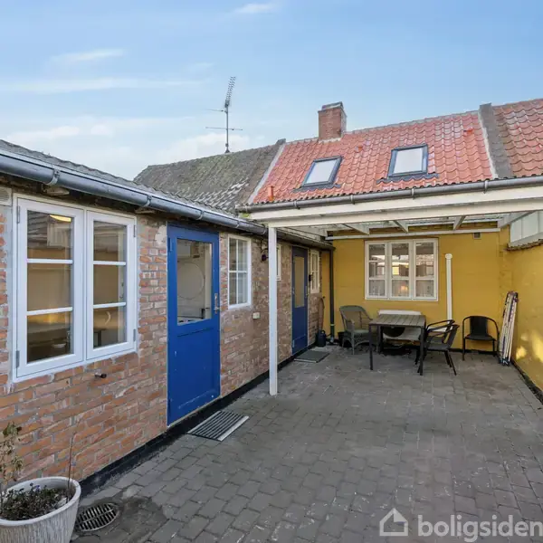 En gårdhave med gule og murstensvægge, blå døre og et halvtag. Bord og stole står ovenpå flisebelægning, omgivet af farverige terrassevægge og plantekrukker.