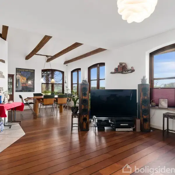 Brændeovn opvarmer en stue med synlige bjælker og trægulv, mens en fladskærm står centralt. Flere store vinduer lukker lys ind; spisebordet står ved siden af.