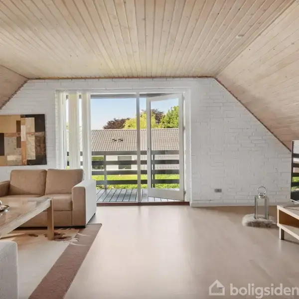 Sofa står i en lys stue med skråloft, der åbner ud mod en balkon med glasdøre. Ved siden af sofaen ses et sofabord og et TV på et lavt skab.