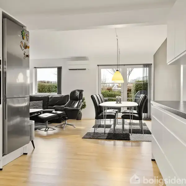 Et åbent køkken-alrum med hvidt køkken, stål-køleskab, og spisebord med sorte stole i centrum. Store vinduer viser en have udenfor, skaber et lyst og rummeligt miljø.