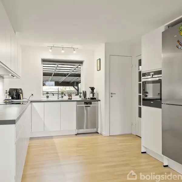 Køkken med hvide skabe og sort bordplade. Køleskab med magneter står ved siden af en indbygget ovn. Trægulv og hvidmalede vægge skaber en lys atmosfære.