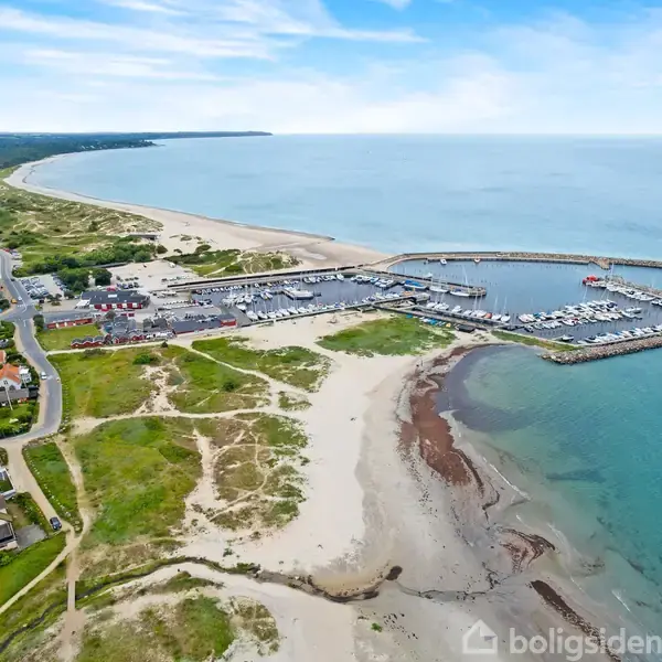 Havn med både ligger stille ved kysten, omgivet af strande og grønne arealer. Til venstre er der en by med røde hustage. Horizonten viser en stor bugt.