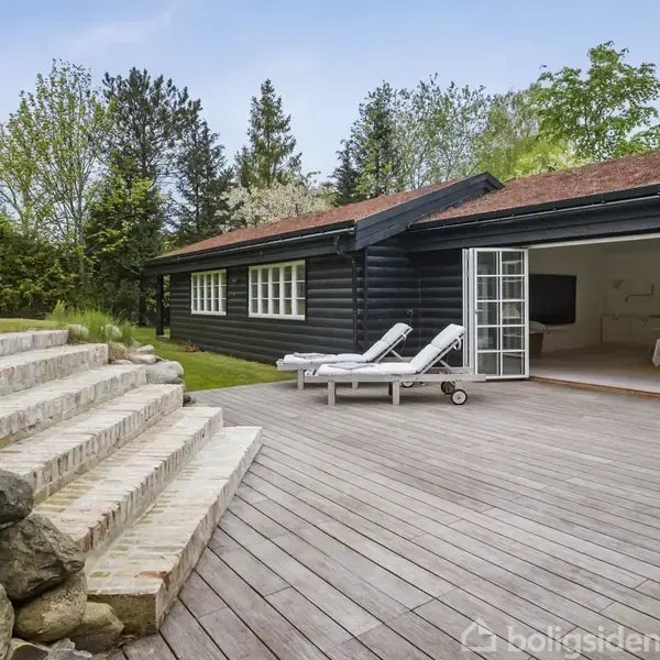 En sort træhytte med åbne døre fører til en terrasse med liggestole og piknikbord. Stenbelagt trappe stiger fra terrassen. Omgivet af frodige træer og buske.