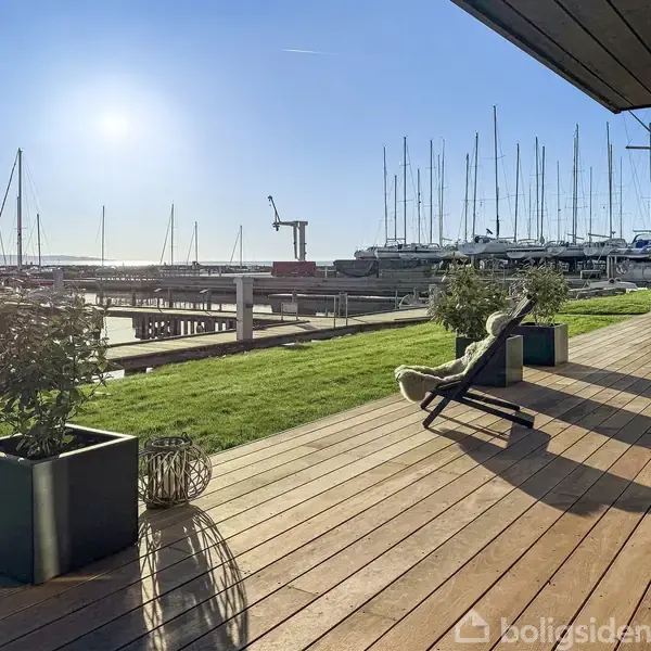 Terrasse med trædæk med solstole og plantekrukker, ser ud over en marina med sejlskibe. Solen skinner klart på himlen, og der er grønne græsarealer i nærheden.