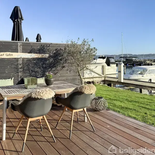 Et træbord med to stole står på en terrasse med udsigt over en marina. Solen skinner. Teksten "Lazy Lounge" ses på en væg.