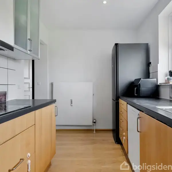 Køkken med lyse træskabe og mørke bordplader. Et køleskab står i hjørnet. En vask ved vinduet med udsigt til en blå himmel udenfor. Der er ingen tekst.