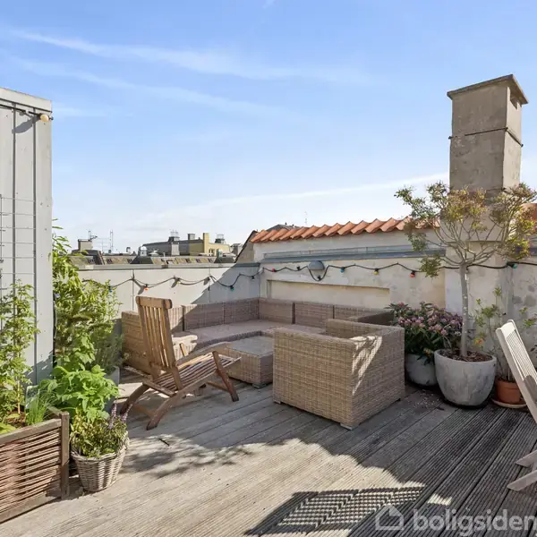 Terrasse indrettet med flettede sofaer og stole ved et træbord, omkranset af planter i potter. Solskinsdag med klar blå himmel og udsigt over byens tage.