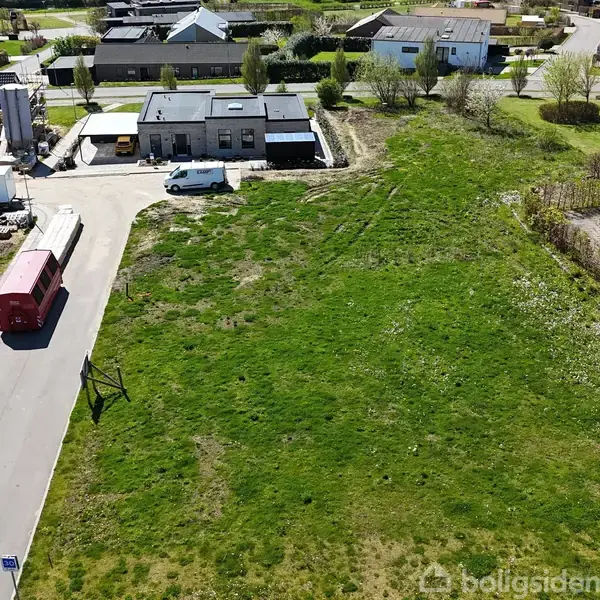 Grønt græsareal ved siden af en vej med en rød bus parket. I baggrunden er der boliger og parkerede biler i et villakvarter.