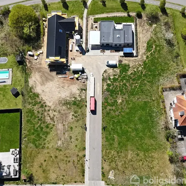 Luftfotografi viser en vej med græsklædte områder på begge sider, omgivet af moderne bygninger og huse med træer og en swimmingpool i kvarteret.