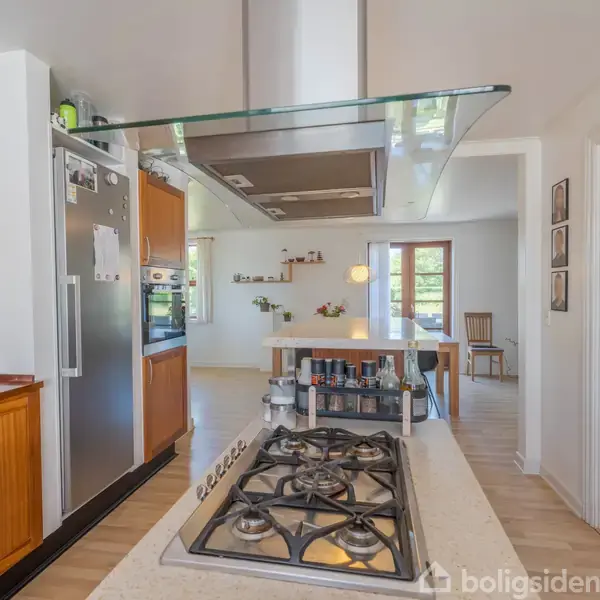 En moderne induktionskogeplade med fem brændere i et køkkenmiljø. Baggrunden viser et spiseområde med bord og stole samt en dørløs åbning mod et lyst rum.