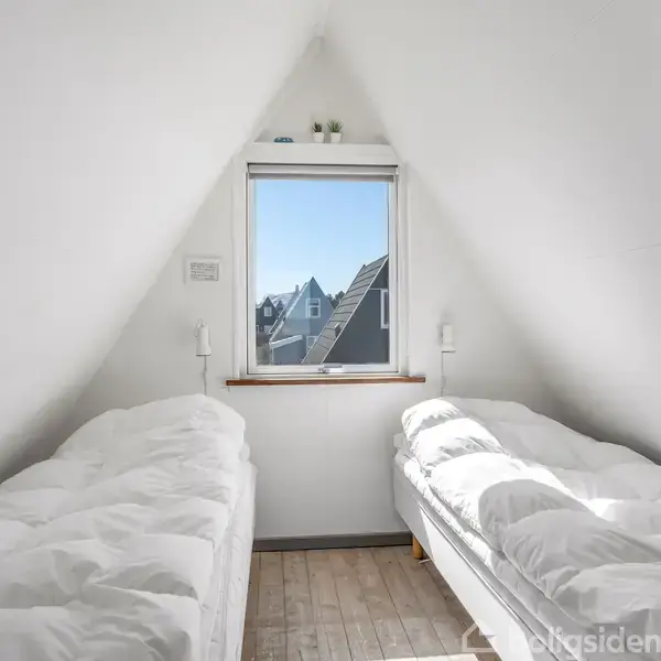 To enkeltsenge med hvide dyner står ved siden af hinanden under et skråt loft. Ét vindue mellem dem viser tage af nærliggende huse. Ingen tekst er synlig.