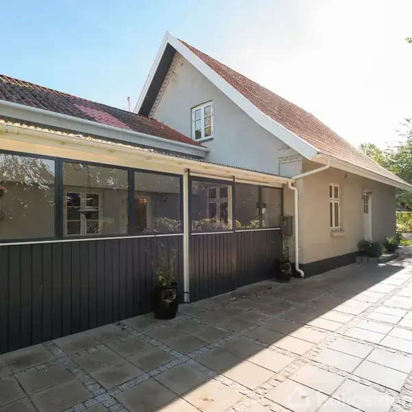 Et hvidt hus med rødtag står i solskin omgivet af flisebelagt terrasse og grønne planter. En glaspanelvæg og flere vinduer ses langs huset.