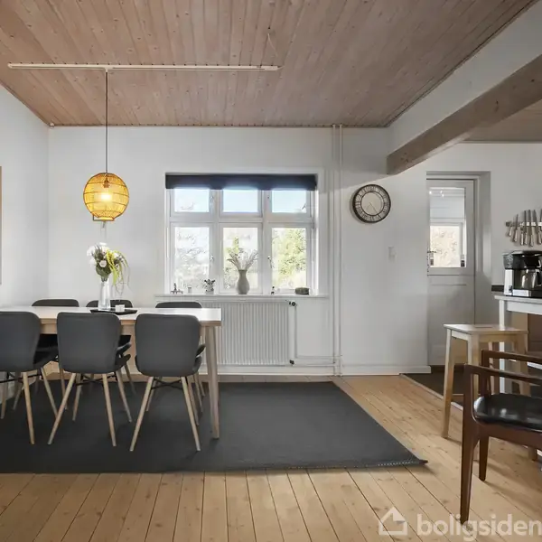 Spisebord med fem stole står i et lyst rum med trægulv og hvidmalede vægge. En lampe hænger over bordet, og en brændeovn er indbygget i væggen.