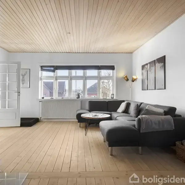 Stue med grå hjørnesofa mod en hvid væg, ved siden af et indbygget brændeovn. Gennem store vinduer strømmer dagslys ind. Trægulve og loft skaber en varm atmosfære.