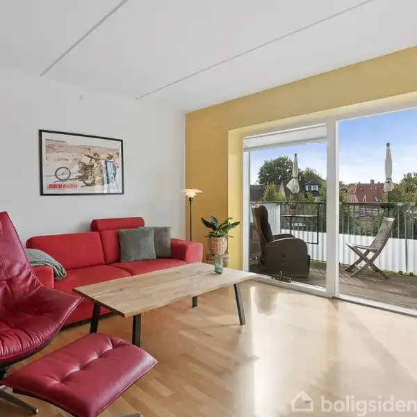 En rød lænestol med fodskammel står ved siden af en rød sofa ved et sofabord i en stue. Gennem glasdøren ses en altan med stole og udsigt til bygninger.