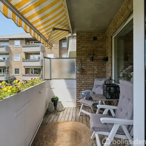 Balkonen har to komfortable stole med hynder placeret under en gul- og hvidstribet markise, omgivet af blomsterkasser. Udendørs er der udsyn til et lejlighedskompleks og grønne områder.