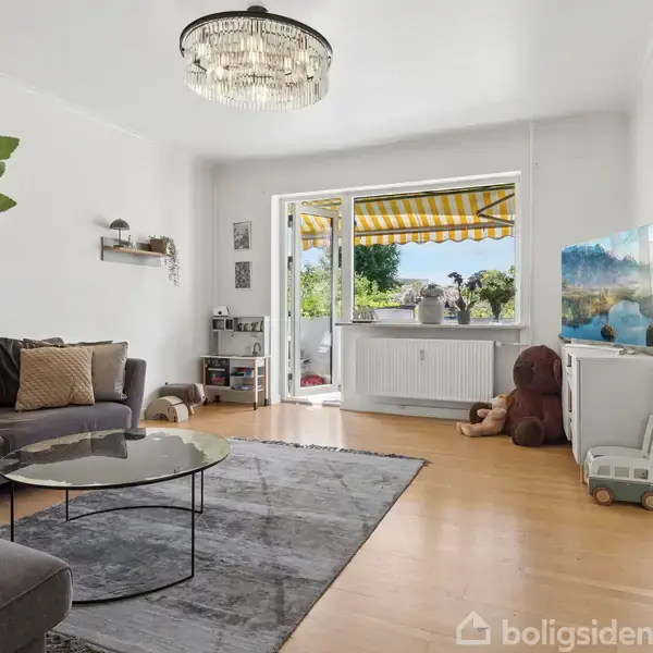 Grå sofa står i et lyst stueområde med moderne indretning. Et stort vindue åbner ud til en terrasse med gul-og-hvid-stribet markise. Et tæppe, planter og en lysekrone.