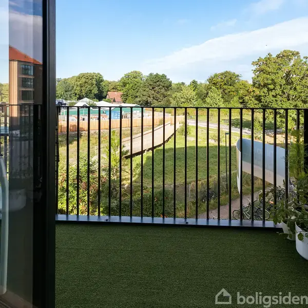 Balkon med grøn kunstgræs og sort metalrækværk. En stol står tæt på glasdøren. I baggrunden ses en park og et landskab med træer.