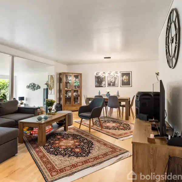 En stue med en grå hjørnesofa og sofabord, flankeret af tæpper. I baggrunden står et spisebord under en lysekrone. Grønne planter dekorerer rummet, der er lyst og moderne.
