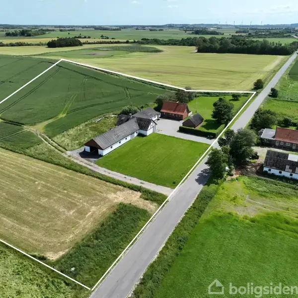 Gård omgivet af marker, kendetegnet ved hovedbygning og udhuse. Det ligger tæt på en vej med træer langs siderne. Landskabet er fladt med spredte træer i horisonten.