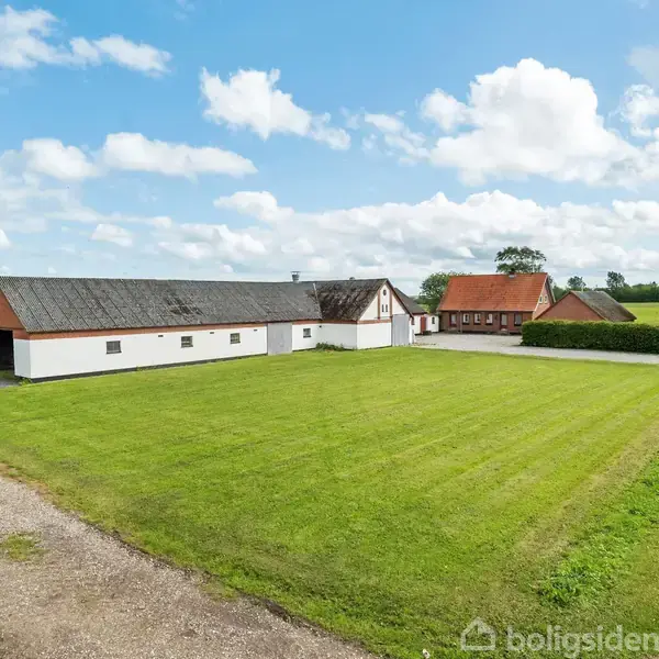 En lang, hvid bygning med rødt tag står ved siden af en græsplæne og en grusvej. I baggrunden ses en anden rød bygning og grønne marker.